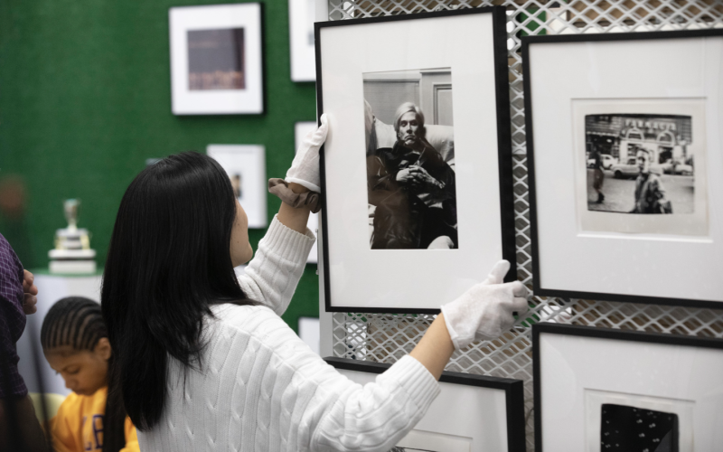 A woman with long black hair in a white sweater and white gloves reaches for a framed black-and-white photo of Andy Warhol that hangs on a wall next to other framed photos.