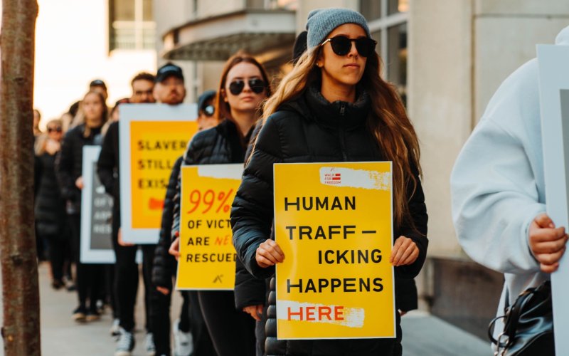 A line of people march, holding yellow anti-trafficking signs. At front is a women in sunglasses with a sign reading "Human Trafficking Happens Here"