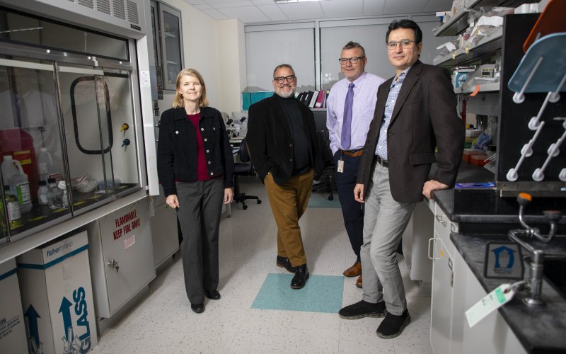 The four featured researchers stand in Melinda Larsen's lab in the RNA Institute. The room itself is dimly lit, with the camera flash adding light. From left to right: Melinda Larsen (woman with blond hair wearing a black jacket with gray pants), Scott Tenenbaum (man with black glasses wearing black jacket with khaki pants), Andre Melendez (man wearing black rimmed glasses, pale purple dress shirt and tie and black slacks, and Mehmet Yigit (man wearing wire rimmed glasses, brown jacket and grey pants).
