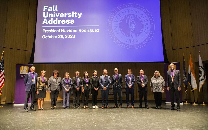 A line of 13 people standing shoulder to shoulder on a stage in front of a blue screen that reads: "Fall University Address, President Havidan Rodriguez, October 26, 2023."