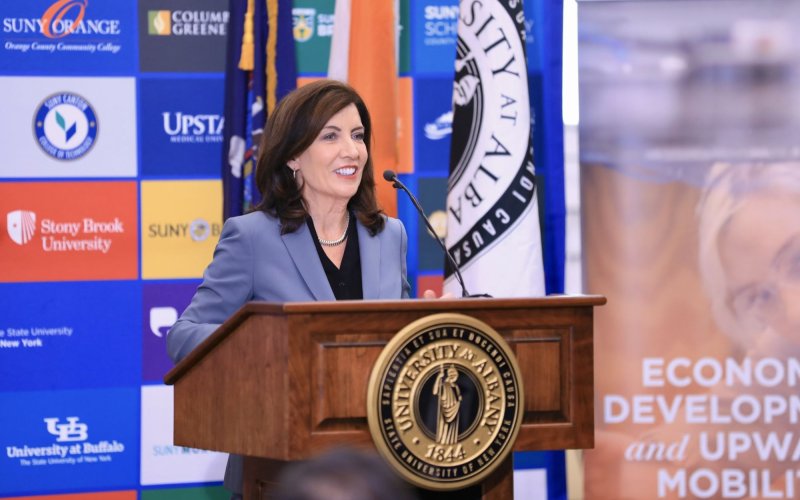 Gov. Hochul speaks from a wooden podium with the University at Albany seal, in front of a colorful poster of SUNY college logos