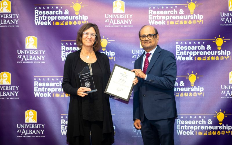 Barbara Sutton stands right dressed in black, with an award in one hand while accepting a certificate from Thenkurussi (Kesh) Kesavadas. Kesh stands on the left, presenting the certificate in a blue suit. Behind them is a repeating backdrop with repeating logos that reads "Research & Entrepreneurship Week" and "University at Albany".
