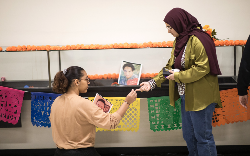 A young woman with a salmon colored shirt, ponytail and glasses kneels on the floor in front of a glass display case decorated with colorful garlands and pictures of deceased loved ones. She accepts a piece of tape from a young woman with a burgundy head scarf and green blouse who stands above her.