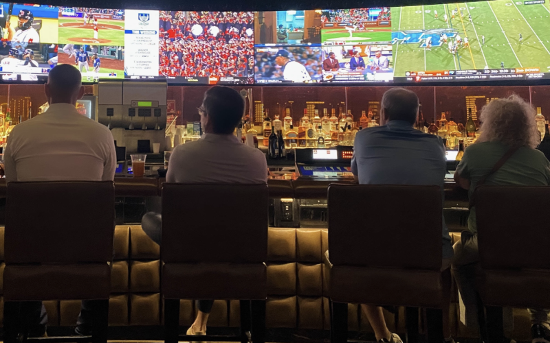 Four people are seated at a dimly-lit bar watching sports on a row of TV screens