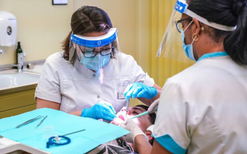 Two dental health care workers wearing white scrubs, blue gloves, face masks, face shields and eye glasses conduct a procedure on a patient using dental implements.