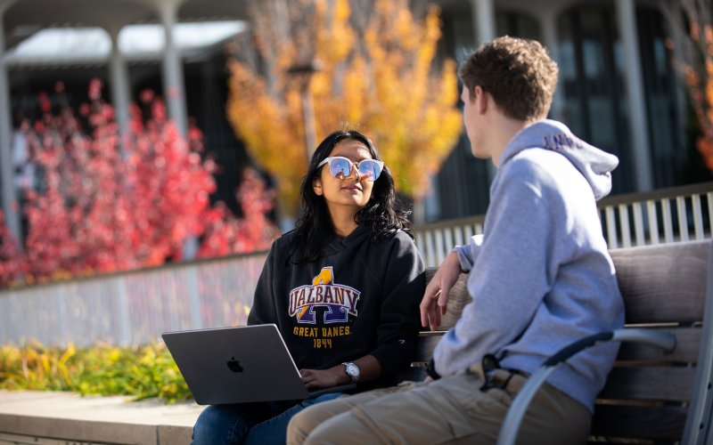 Two students wearing sweatshirts sit on a bench on a sunny fall day. One holds a laptop on their lap.