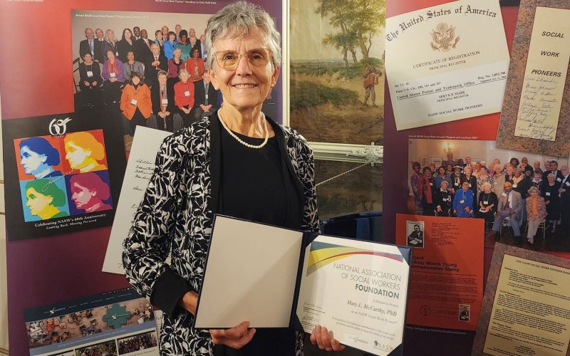 A smiling Mary McCarthy holds her award while standing in front of a panel with photos and posters 