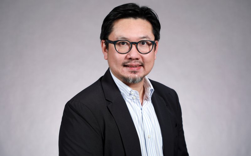 Portrait of Ricky Leung wearing glasses, a black blazer and a white, striped button-down shirt, seated in front of a gray background.