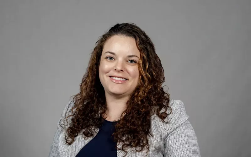 Sarah Domoff poses for a portrait against a gray background wearing a light gray jacket over a navy blue shirt. Domoff is smiling and has brown curly hair. 