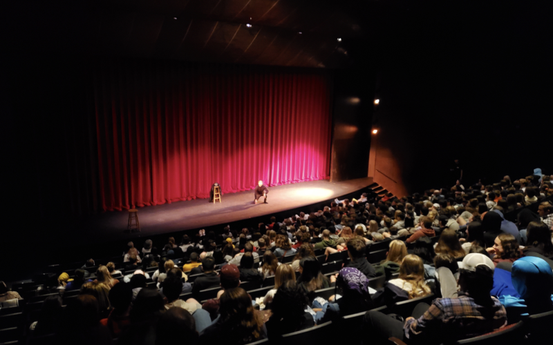 An auditorium full of high school students watches a man dressed in black kneeling on stage in front of a red curtain at the UAlbany Performing Arts Center.