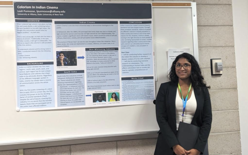 A woman in a blazer and glasses holds a folder and stands in front of a research poster titled "Colorism in Indian Cinema."