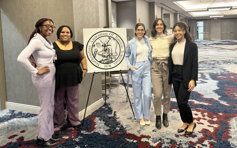 Five women dressed in business attire students pose and smile outside of a conference room on either side of a sign that reads, "Northeastern Association Of Forensic Scientists 1976"