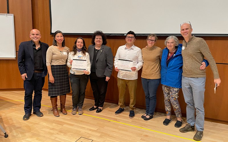 From left to right,  Andrew Poulos, Cheryl Andam, Manuela Montoya Giraldo, Jeanette Altarriba, Andrew Munoz Gamba, Cara Pager, Marlene Belfort and Ken Halvorsen pose and smile in a conference room.  Manuela and Andrew both hold certificates. 