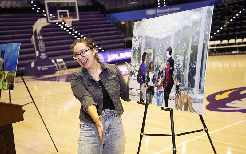 A woman stands on a basketball court smiling and gesturing toward an easel with a digital drawing of students standing in the snow on the UAlbany campus with a leashed Great Dane.