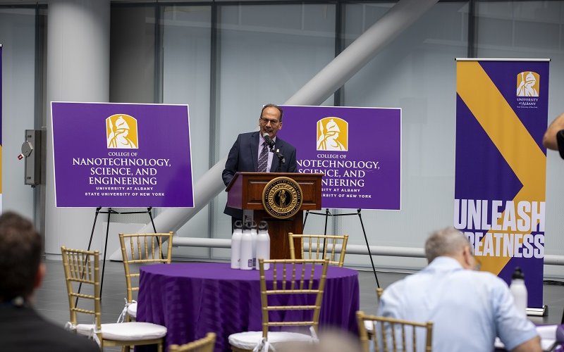 A man speaks from a brown lectern in front of purple tables and flanked by rectangular purple signs that read: "College of Nanotechnology, Science, and Engineering"