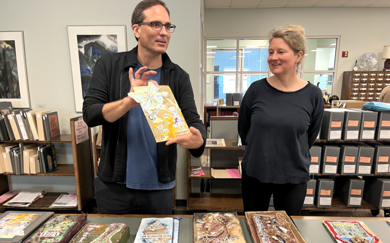 A man with short black hair and glasses holds a handmade art book while a woman with gray hair watches. They stand in front of a table topped with handmade books.