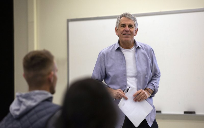 A gray-haired professor stands in front of a class holding his glasses and some papers.