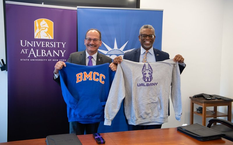 Two men stand smiling hold up sweatshirts from each other's colleges. One is blue and says "BMCC," while the other is gray with a purple Great Dane face and says "UAlbany."