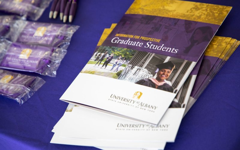 A pile of brochures reading "Information for prospective Graduate Students, University at Albany" on a purple tablecloth