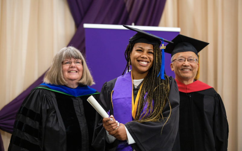 A smiling graduate in a black cap and gown holds a diploma as she poses for a portrait on stage with two others dressed in black graduation robes.