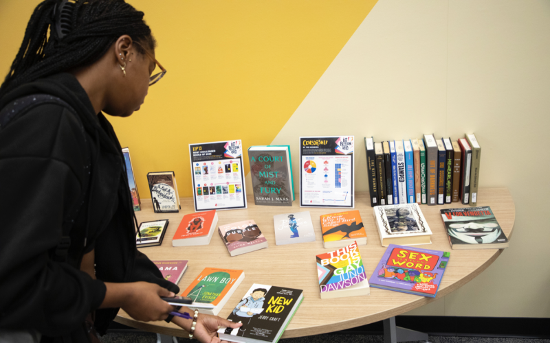 A young woman with long black braids and glasses picks up a book, titled "New Kid" off of a table topped with a diverse range of books on display.