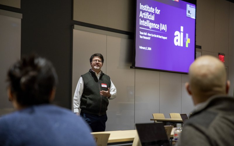 Eric Stern stands under a projection screen with the words “Institute for Artificial Intelligence” as he speaks to people seated in front of him.