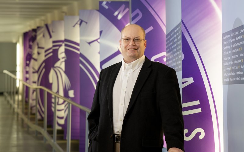 Jesse Holland, MBA 2023, stands in a hallway in front of a series of wall hangings that showcase the seal of the University at albany in puple and white.