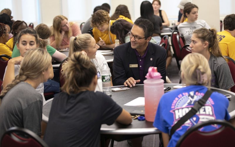 A man in glasses and a black jacket listens, smiling, to a student talking to him at one of many round tables in a room full of students talking and writing.