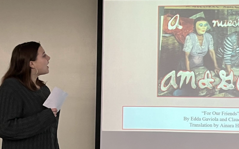 A young woman with shoulder length brown hair and hoop earrings speaks at the front of a room next to a screen depicting a woman in a dress and plastic face mask.