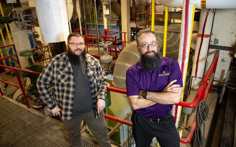 Two men with beards stand on a catwalk in an industrial setting in front of two stories of massive boiler equipment.