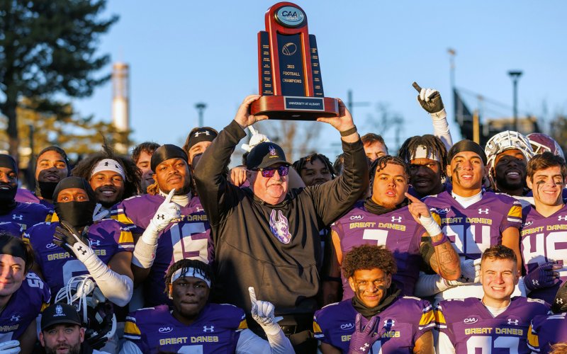 UAlbany head football coach Greg Gattuso hoists the CAA Championship trophy, surrounded by the team, following the end of the 2023 regular season
