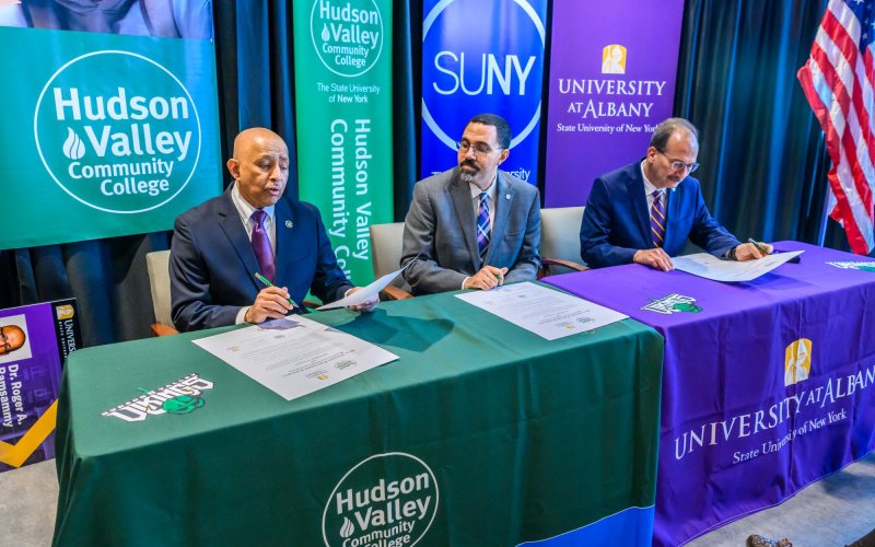 Three men in suits sign oversized pieces of paper at a table with a gree Hudson Valley Community College tablecloth on one side and a purple University at Albany tablecloth on the other. HVCC, SUNY and UAlbany banners are behind them.