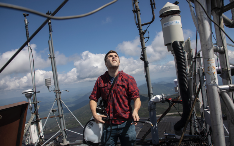 A man in a red checkered shirt stands at the top of UAlbany's Atmospheric Sciences Research Center Whiteface Mountain Summit Weather Station. He is holding a piece of equipment and squinting at the sun. Cloud-topped mountains can be seen in the distance.