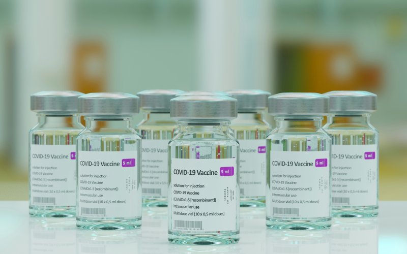 Image features seven COVID-19 vaccine vials with silver lids, arranged on a sleek, white tabletop. The background is blurred.