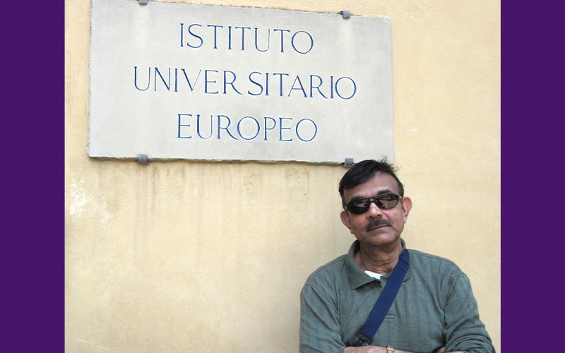 Kajal Lahiri stands with his arms crossed wearing a green long sleeved shirt and sunglasses. A blue bag strap falls across his chest and the bottom of the sign on the wall behind him reads "Instituto Universitario Europeo."