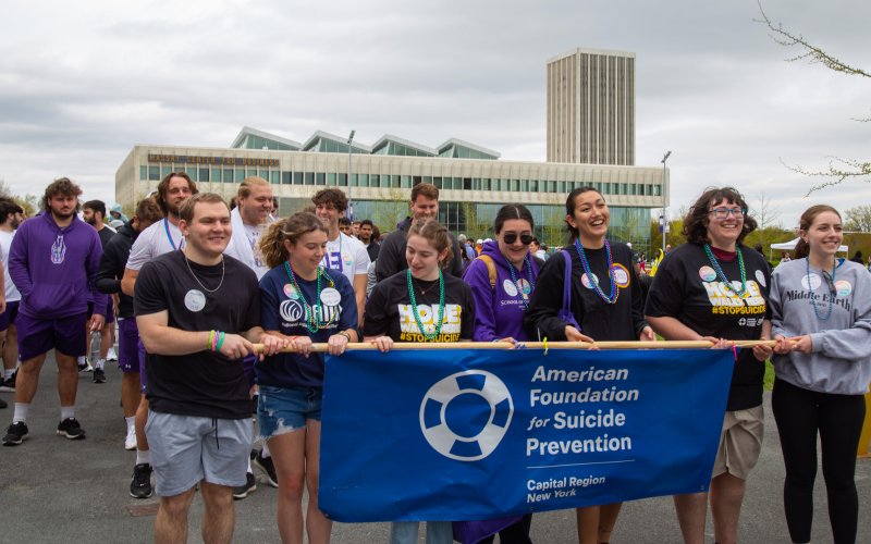 Participants line up for the 2023 Out of the Darkness Walk at UAlbany’s Entry Plaza. In front, seven smiling students hold a blue banner suspended on a wooden rod that says: "American Foundation for Suicide Prevention, Capital Region New York". The sky is cloudy and the Massry Business School can be seen in the background. 