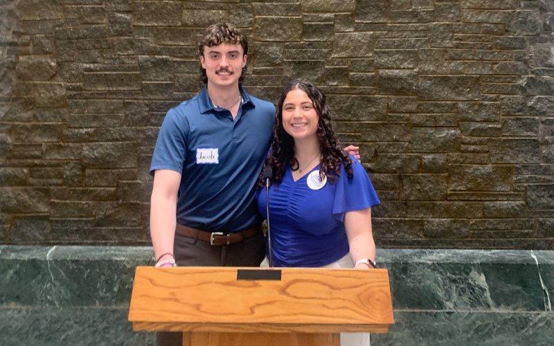 Jacob Schoff and Alexis Candodo pose for a photo behind a wooden podium in the NYS Capitol building. Both are smiling and wearing blue short sleeved shirts. The wall behind them is made of brown stone and green marble.