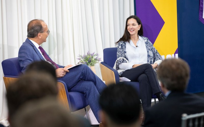 A man and woman sit on chairs in conversation, in front of an audience.