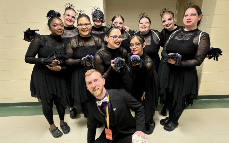 A man dressed in all black and a bowtie kneels in front of a group of young women wearing black dresses, spider web crowns and holding stuffed animal spiders.