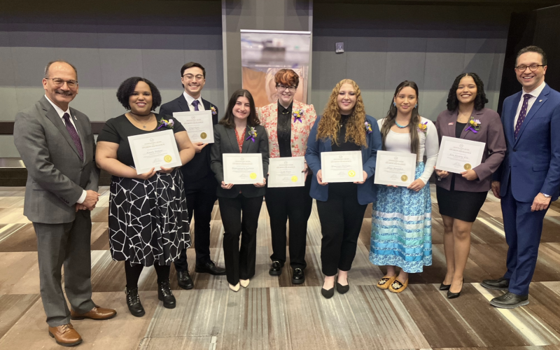 Seven students in professional attire stand holding award certificates and smile for a picture with UAlbany President Havidán Rodríguez and Vice President for Student Affairs Michael Christakis in a conference room.