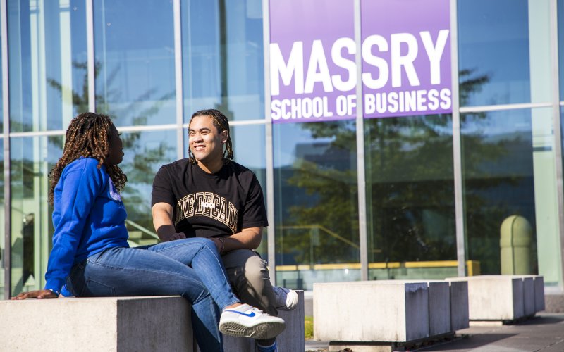 Two students sit outside in front of a glass window with a purple sign that says Massry School of Business.