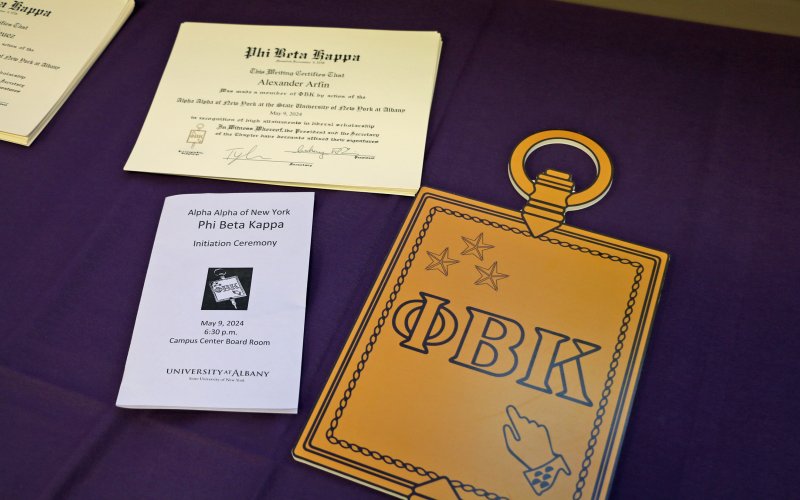 UAlbany Phi Beta Kappa programs and award certificates are on a table with a purple background.