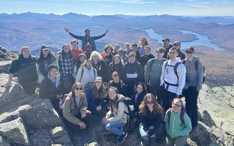 An undergraduate class stands atop Whiteface Mountain with fall foliage behind them.