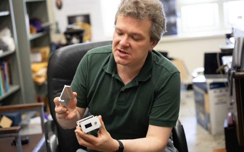A a seated man in a green polo shirt leans toward the camera while holding a small, rectangular metallic box.