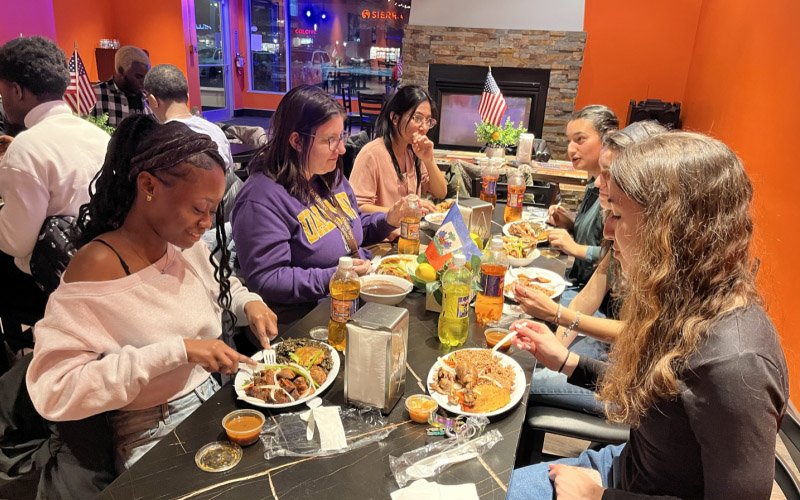Students are seated at a table in a restaurant eating plates of Haitian food.