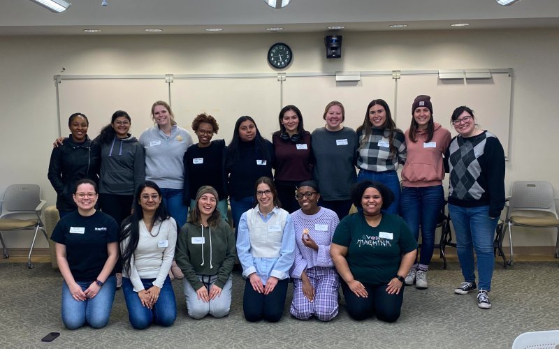 A group of 16 members of STEM NOW pose, all smiling, for a group photo in a classroom at UAlbany. The group is posed in front of a white board and they are arranged in two rows. The front row is kneeling on the carpet, which is gray. All are wearing white name tags.
