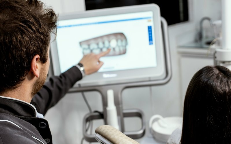This image is of two people in a dentist office, taken from behind. A man with short brown hair points to a digital screen showing an image of a set of teeth. A person with long hair is seated behind him. 
