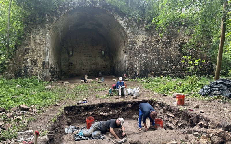 Archaeologists work in a hole digging with tools, with one person recording notes, in the background is an archway of a ruined church from the 1500s.