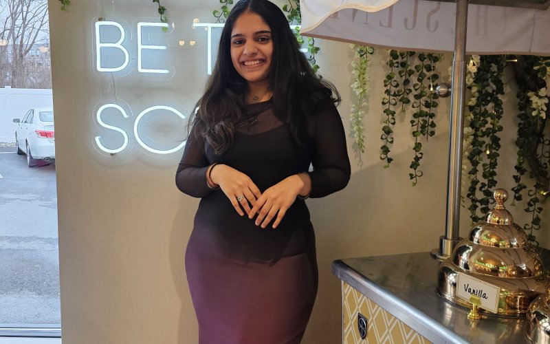 Smiling woman in a black and purple dress stands beside a vintage coffee cart. Neon sign and green vines hang on the wall, creating a cozy ambiance.