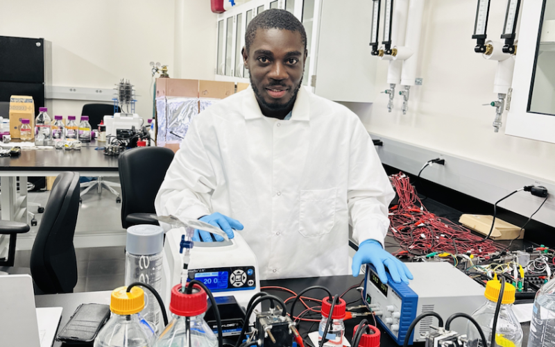 A man with short black hair and a beard wears a white lab coat and blue glove and touches equipment in a laboratory.